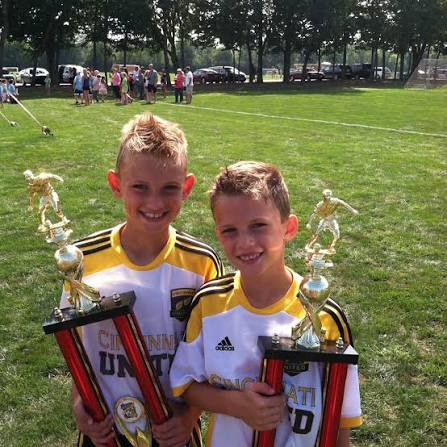 Two young boys in soccer jerseys holding trophies on a field